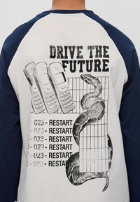Raglan-Ärmelshirt mit einem cremefarbenen Körper und blauen Ärmeln, mit einem Grafikdesign von Mobiltelefonen und einer Schlange, der Text lautet "FAHRE IN DIE ZUKUNFT."
