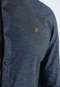 Camisa azul oscuro abotonada, fabricada con tela texturizada, con un pequeño logo dorado en el lado izquierdo del pecho y botones negros en la parte delantera.