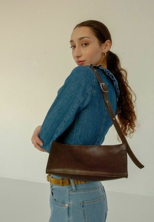 Borsa a spalla in pelle marrone con una superficie liscia, forma rettangolare e tracolla regolabile; indossata con una giacca di jeans blu.