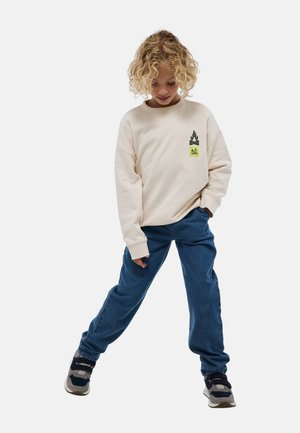 Sweatshirt beige clair avec un graphisme vert sur la poitrine gauche, associé à un jean en denim bleu ample et des baskets noires avec des accents gris.