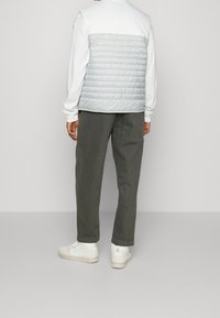 Man i vit långärmad skjorta, ljusgrå vadderad väst, mörkgrå byxor med bakficka och vita sneakers, som står med ryggen vänd.