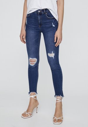 Femme portant un jean skinny bleu foncé déchiré et des sandales blanches à talons hauts à lanières, debout devant un fond uni.