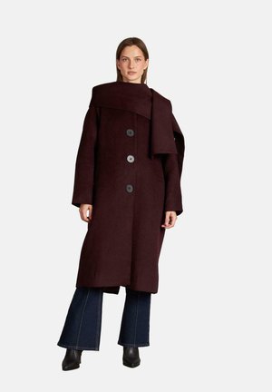 Abrigo de lana burdeos con un amplio collar, presenta tres grandes botones y un diseño envolvente. Combinado con pantalones de mezclilla acampanados y botines.