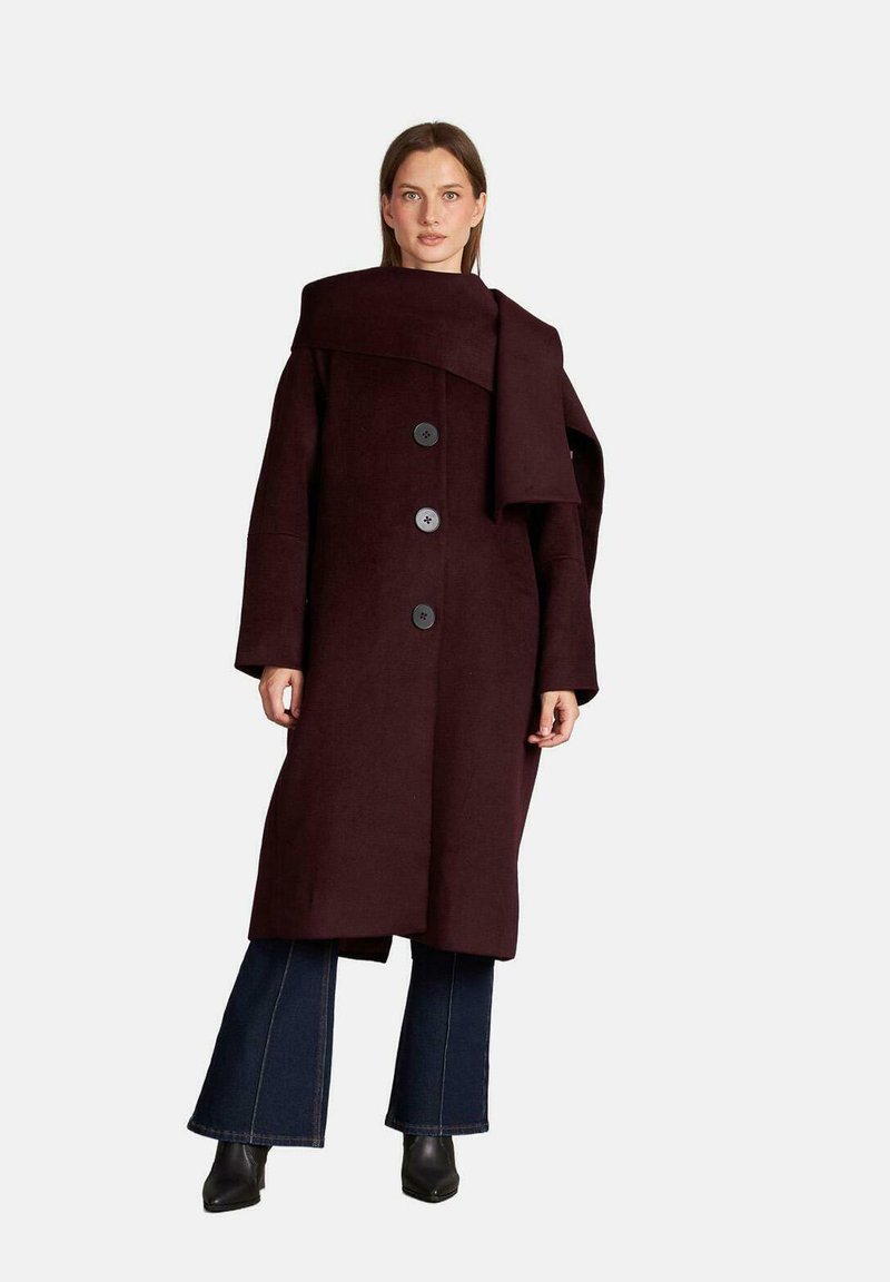 Bordeaux wollen jas met een brede kraag, voorzien van drie grote knopen en een wikkelontwerp. Gecombineerd met uitlopende spijkerbroek en enkellaarsjes.