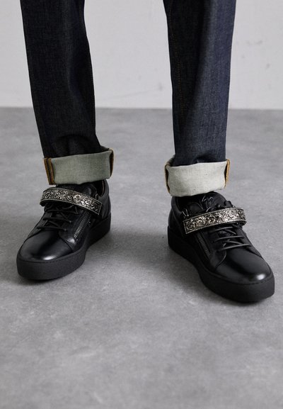 Black leather sneakers with a thick black sole, decorative strap featuring an intricate silver design, paired with dark denim jeans.
