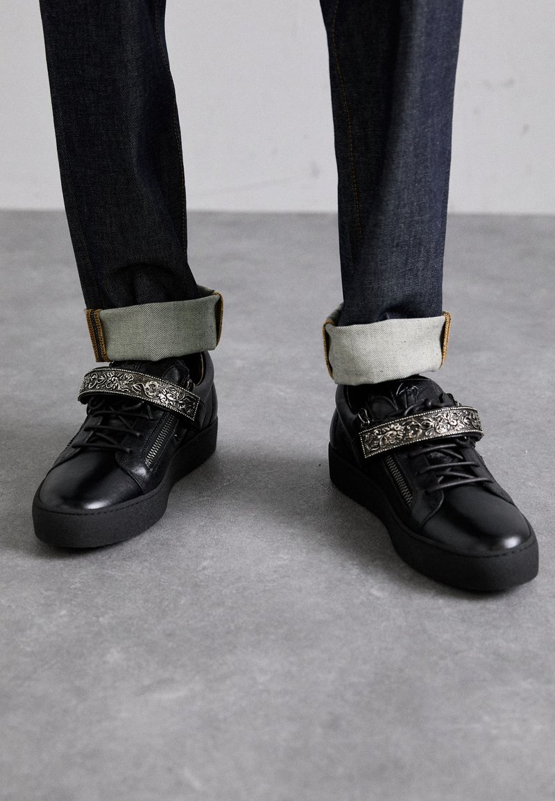 Juodi odiniai sportbačiai su storu juodu padu, dekoratyvine juosta, kurioje yra sudėtingas sidabrinis raštas, derinami su tamsiai džinsais.