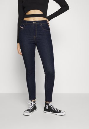 Kvinde iført mørkeblå skinny jeans, sort crop long-sleeve top og sorte høje sneakers står mod en hvid baggrund.