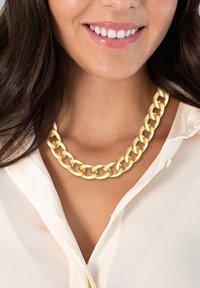 Zlaten kovinski verižni obesek s velikimi prepletenimi povezavami, nosi se čez svetlo obarvano srajco s kratkimi rokavi. Gladka tekstura z sijočim zaključkom.