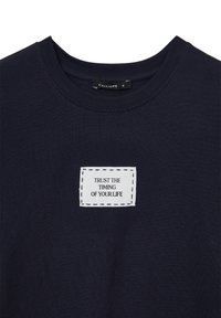 Maglietta a maniche corte blu navy con un'etichetta rettangolare bianca centrata sul petto che recita "FIDATI DEL TEMPO DELLA TUA VITA."