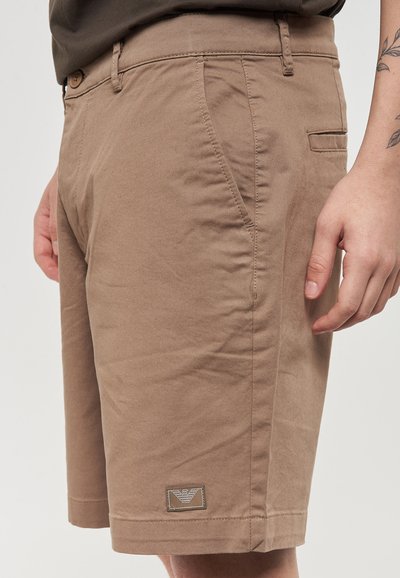 Pantaloni corti in cotone beige con vestibilità dritta, tasche laterali e una tasca posteriore. Presenta un piccolo patch con logo sull'orlo sinistro.