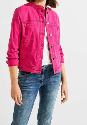 Femme portant une veste boutonnée rose vif sur un haut blanc avec un jean bleu, debout devant un fond clair uni.