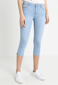 Jeans capri en denim bleu clair avec une coupe slim, de longueur cheville et des fentes latérales à l'ourlet. Associés à des baskets blanches.