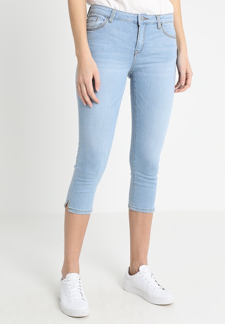 Jeans capri en denim bleu clair avec une coupe slim, de longueur cheville et des fentes latérales à l'ourlet. Associés à des baskets blanches.