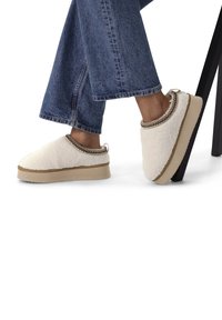 Witte fleeceslippers met een gestructureerd oppervlak en contrasterende bruine stiksels. Dikke beige zool biedt extra hoogte. Denim broek zichtbaar.