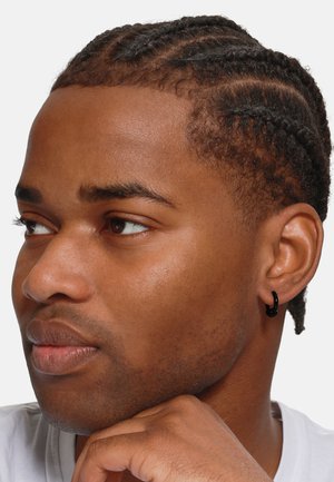 Portrait d'une personne avec des cheveux tressés, portant une boucle d'oreille noire. Affiche un teint moyen et lisse et une expression neutre.