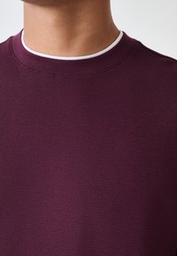 Bordeauxfarbener, strukturierter Sweatshirt mit Rundhalsausschnitt und glattem weißem Kragenabschluss. Nahaufnahme zeigt detaillierten Stoff und Naht.