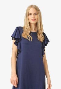 Tamsiai mėlyna suknelė su pūkuotomis trumpomis rankovėmis, švelni tekstūra, apvaliu iškirpimu ir atsipalaidavusiu siluetu, demonstruojanti elegantišką dizainą ir plazdančią formą.