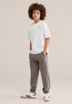 Giovane con capelli ricci in piedi con le mani nelle tasche, indossa una maglietta bianca con stampa grafica, pantaloni jogger grigi e sneaker bianche con strisce nere.