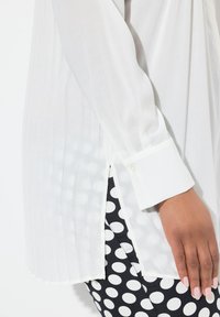 Witte blouse met lange mouwen, verticale plooien, manchetten en knoopsluiting. Gekoesterd met zwarte broek met stippen in de taille.