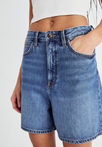 Shorts en denim bleu à taille haute, avec une fermeture à bouton, des poches avant et des détails de coutures visibles. Tissu texturé avec un léger dégradé.