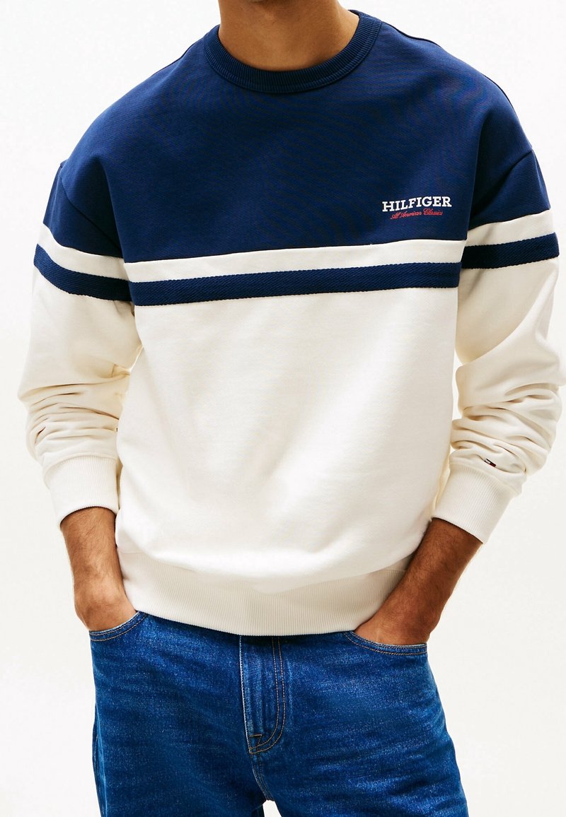 Homme portant un sweat Hilfiger à rayures bleu marine et blanc, les mains dans les poches d'un jean bleu, vu du cou à la taille.