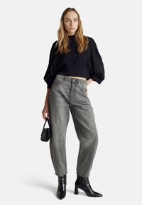 Černý krátký svetr, šedé džíny s vysokým pasem a černé lakované kozačky na kotníky. Modelka drží malou černou kabelku s texturou.