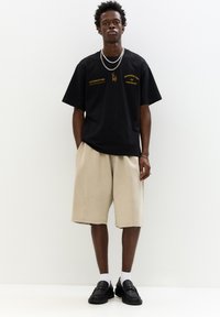 Fekete, túlméretezett, rövid ujjú póló aranyszínű felirattal, bézs, térd feletti sorttal és fekete mokaszin cipővel fehér zoknival párosítva.