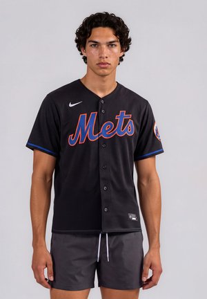 Giovane uomo con capelli ricci indossa una maglia da baseball nera dei Mets e pantaloncini grigi, in piedi davanti a uno sfondo chiaro e uniforme.