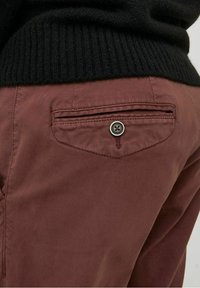 Gros plan sur la poche arrière d'un pantalon bordeaux avec fermeture à bouton, et le bas d'un pull côtelé noir visible au-dessus.