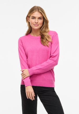 Tânără cu părul lung și blond, purtând un pulover roz intens și pantaloni negri, stând în picioare pe un fundal alb simplu.