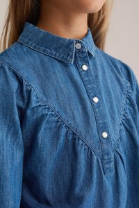 Denim krekls ar apkakles kaklu, ar pogu aizdari priekšpusē, savākta apdare un gaiši zila krāsa ar teksturētu audumu.