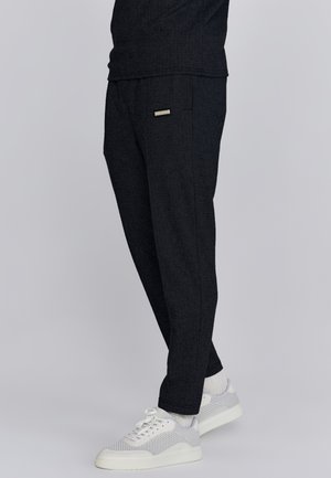 Pantalones de espiga en gris oscuro con corte ajustado, que presentan una etiqueta de marca en el bolsillo delantero y se combinan con zapatillas gris claro.