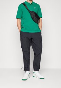 Man med grön Nike pikétröja, svarta cargobyxor, vit och grön sneakers, och svart axelremsväska mot en enfärgad bakgrund.