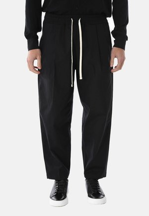 Uomo che indossa pantaloni neri larghi con coulisse bianca e sneakers nere con suole bianche, in piedi contro uno sfondo semplice.