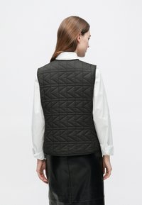 Črni prešiti telovnik z diamantnim vzorcem, sproščenega kroja in brez vidnih zapenjanj, kombiniran z belo srajco z dolgimi rokavi.