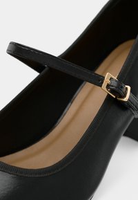 Čierne topánky typu Mary Jane so zlatou prackou a svetlohnedou vnútornou vložkou, na ktorých sú vidieť remienok a časť otvoru topánky.