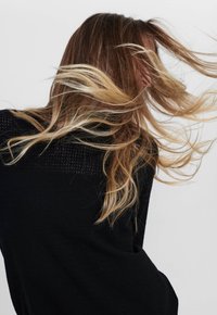 Femme aux longs cheveux ondulés blond et brun, portant un haut noir, les cheveux flottant comme pris en mouvement sur un fond clair uni.