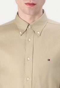 Beige gombos ing, pamutanyagból készült, klasszikus gallérral, gombos elülső résszel és egy kis hímzett logóval piros, fehér és kék színekben.