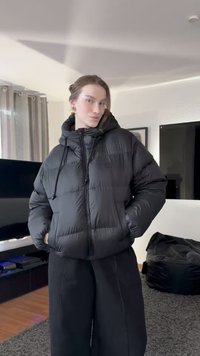 Črna puhasta jakna s kapuco, ima zadrgo, vrvice in prešite vzorce. Model pozira z rokami v žepih.