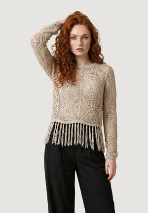 Giovane donna con capelli rossi ricci che indossa un maglione beige all'uncinetto con orlo a frange e pantaloni neri a gamba larga, in piedi davanti a uno sfondo semplice.