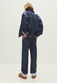 Persoon met blond haar, gekleed in een donkerdenimjack, bijpassende jeans met boorden en bruin instapschoenen, staand tegen een effen achtergrond.