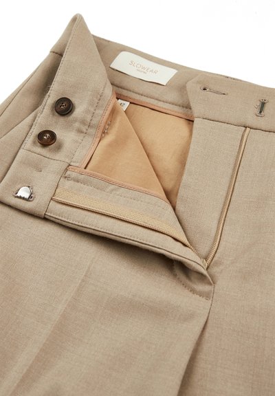 Beige broek met een gladde textuur, voorzien van bruine knopen, een ritssluiting en een tan binnenvoering. Het label zegt "SLOWEAR."