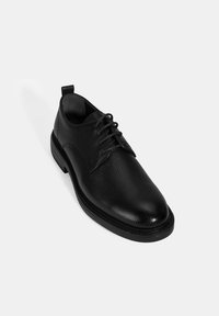 Zwarte leren veterschoen met een gestructureerde afwerking, ronde neus en een contrasterende gladde zool. Voorzien van een trekband aan de achterkant.