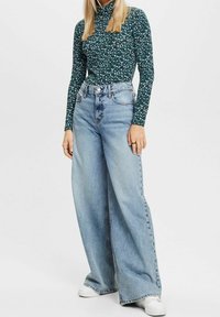 Top floreale a maniche lunghe, verde scuro, abbinato a jeans leggeri a vita alta e a gamba larga, blu chiaro, e sneaker bianche. Tessuto morbido con una vestibilità comoda.