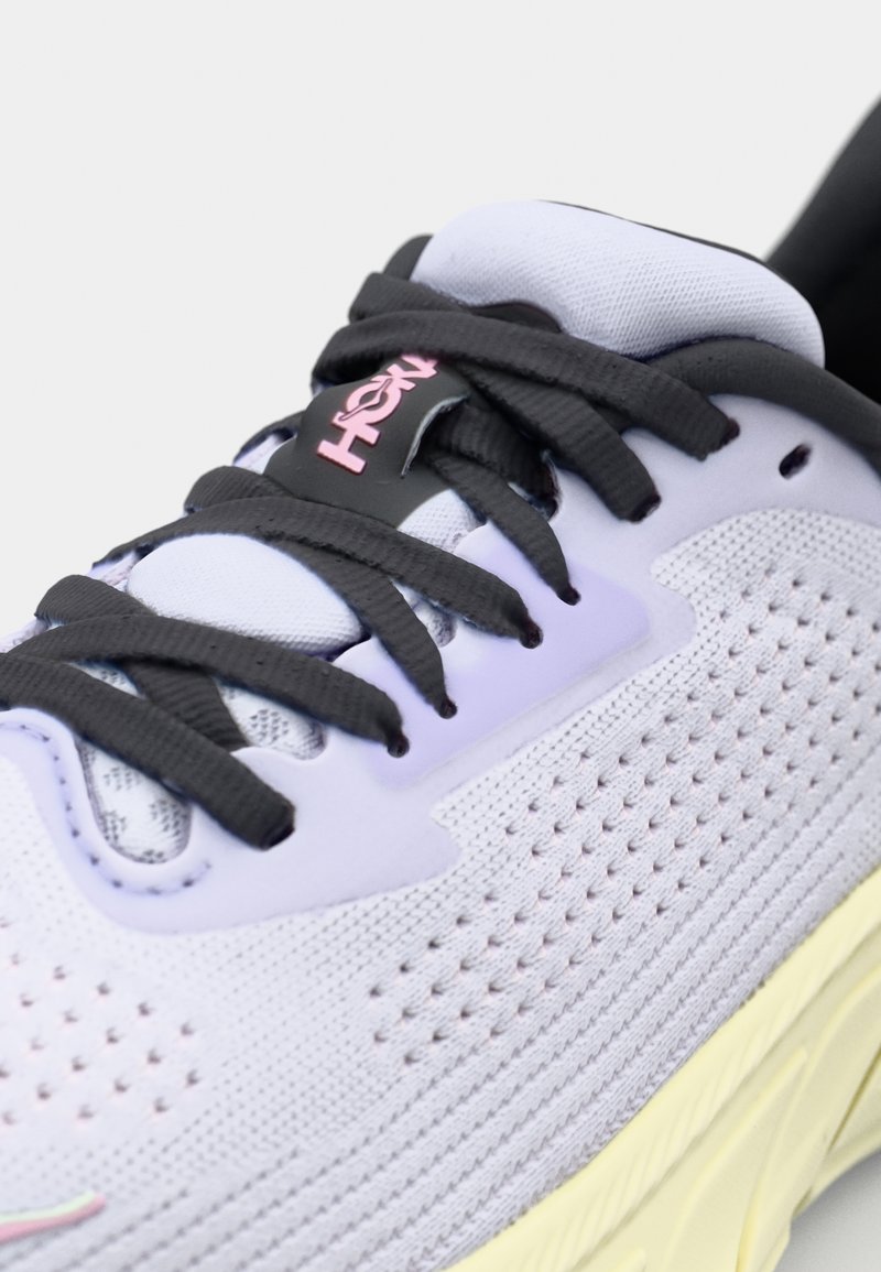 Zapato deportivo de color púrpura claro con cordones negros, parte superior de malla, detalles perforados y suela amarilla. Presenta un logo rosa en la lengüeta.