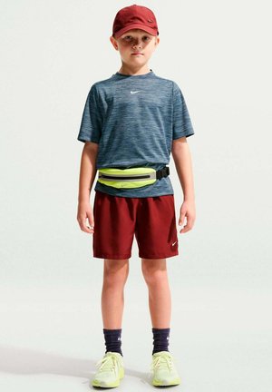 Jongen met een rode pet en korte broek, een blauw Nike-shirt, zwarte sokken, gele hardloopschoenen en een neongele heuptas, staand tegen een witte achtergrond.