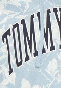 Felpa blu chiaro con motivi floreali bianchi, grande testo "TOMMY" in blu navy scuro, con cappuccio e texture morbida.