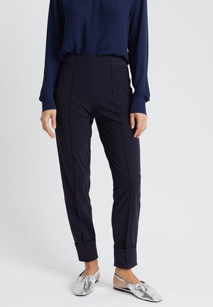Pantalon marine à coupe ajustée, présentant des coutures verticales et des ourlets à revers. Associé à des chaussures plates en métal argenté.