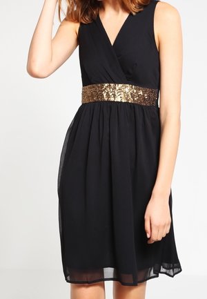 Frau trägt ärmelloses schwarzes Kleid mit tiefem V-Ausschnitt und breitem goldfarbenem Pailletten-Gürtel in der Taille, steht vor einfarbigem Hintergrund.