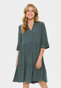 Femeie zâmbitoare cu păr lung blond, îmbrăcată într-o rochie lejeră de culoare verde pădure, cu mâneci evazate până la cot.